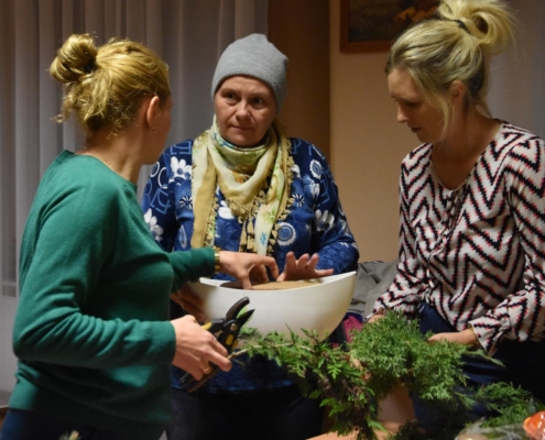 Instruktorka warsztatów, ubrana w białą bluzkę w kolorowe paski, wraz z dwiema uczestniczkami wykonują kompozycje z gałązek tuji, które trzyma instruktorka. Jedna z pań, ubrana w zielony sweter trzyma w dłoni sekator, druga ubrana w niebieską bluzkę w białe kwiaty, czapkę oraz chustkę, pomaga ułóżyć w białym naczyniu gąbkę florystyczną.