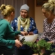 Instruktorka warsztatów, ubrana w białą bluzkę w kolorowe paski, wraz z dwiema uczestniczkami wykonują kompozycje z gałązek tuji, które trzyma instruktorka. Jedna z pań, ubrana w zielony sweter trzyma w dłoni sekator, druga ubrana w niebieską bluzkę w białe kwiaty, czapkę oraz chustkę, pomaga ułóżyć w białym naczyniu gąbkę florystyczną.