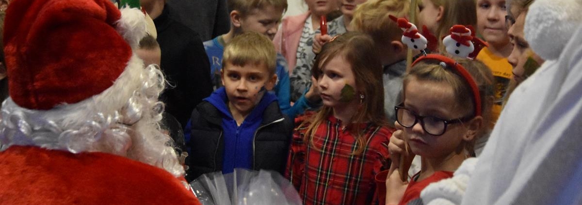 Gromada dzieci zebrana wokół świętego Mikołaja czeka zniecierpliwiona na prezenty. Niektóre z dzieci mają na głowach świąteczno-zimowe opaski. Jedna dziewczynek ma na policzku wymalowaną choinkę.
