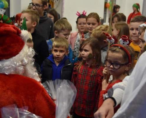 Gromada dzieci zebrana wokół świętego Mikołaja czeka zniecierpliwiona na prezenty. Niektóre z dzieci mają na głowach świąteczno-zimowe opaski. Jedna dziewczynek ma na policzku wymalowaną choinkę.