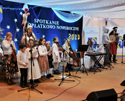 Grupa śpiewających dzieci oraz dorosłych osób - muzyków grających na wiolonczeli, cymbałach, bębenku obręczowym i skrzypcach, stojących przy mikrofonach na tle granatowego materiału dekoracyjnego z napisem "Spotkanie Opłatkowo-Noworoczne"