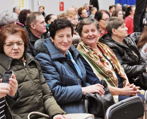 Grupa ludzi siedzących na krzesłach na widowni, dwie kobiety w wieku senioralnym uśmiechają się do obiektywu