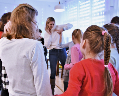 Gromada dzieci stoi w kole, na środku widać instruktorkę GOK, która pokazuje jednemu z uczestników zasady gry. Na pierwszym planie widać dwie dziewczynki obrócone tyłem - po lewej w białej bluzce i długimi włosami, po prawej w czerwonej bluzce i długim warkoczu.