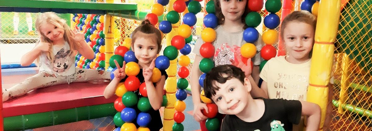 Pięcioro dzieci pozuje do zdjęcia wśród kolorowych, wiszących kulek. Jedno z dzieci - uśmiechnięta dziewczynka w blond włosach siedzi z tyłu na materacach jedną ręką machając a drugą pokazując gest "OK".