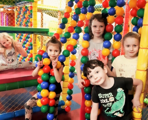 Pięcioro dzieci pozuje do zdjęcia wśród kolorowych, wiszących kulek. Jedno z dzieci - uśmiechnięta dziewczynka w blond włosach siedzi z tyłu na materacach jedną ręką machając a drugą pokazując gest "OK".