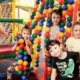 Pięcioro dzieci pozuje do zdjęcia wśród kolorowych, wiszących kulek. Jedno z dzieci - uśmiechnięta dziewczynka w blond włosach siedzi z tyłu na materacach jedną ręką machając a drugą pokazując gest "OK".