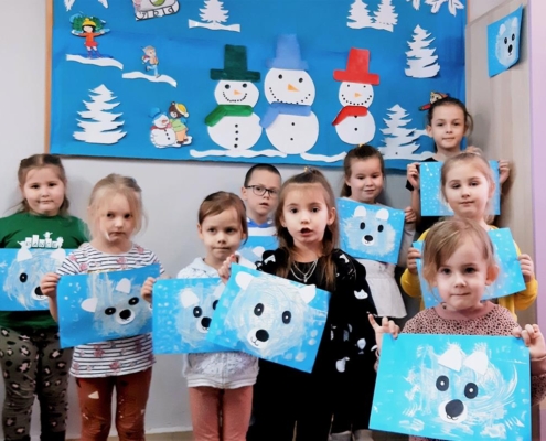 Grupa licząca dziewięcioro dzieci pozuje do zdjęcia. W dłoniach trzymają swoje prace - białe niedżwiadki polarne na niebieskich kartkach. Za dziećmi na tablicy widzimy trzy bałwanki w kolorowych czapkach i szalikach, choinki oraz dzieci bawiące się na śniegu. To wszystko wykonane jest z papieru.