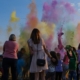 Zdjęcie pokazuje grupę ludzi - dorosłych i dzieci biorących udział w festiwalu kolorów holi. Na zdjęciu został uchwycony moment rzucenia kolorowymi proszkami. Na tle niebieskiego nieba widać kolorowe proszki i chmury powstałe z proszków.