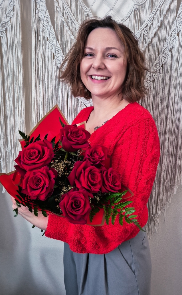Uśmiechnięta kobieta trzyma bukiet czerwonych róż. Ma na sobie czerwony sweter i szare spodnie. Stoi na tle białej makramy. Jej włosy sięgają ramion i są falowane.