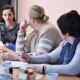 Na zdjęciu widać dwie kobiety zwrócone w stronę koordynatorki projektu, która opowiada o nim. Koordynatorka jest ubrana w szarą bluzkę w falbanki, w białe kwiatki. Ma rozpuszczone ciemne włosy. Na stole widać