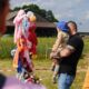 Mężczyzna ubrany w czarną bluzkę z krótkim rękawem i jasne jeansy trzyma na rękach chłopczyka. Chłopczyk ubrany jest w jasnożółtą bluzeczkę ze wzorkami - drzewkami i zwierzątkami. Ma materiałowe zielone spodnie i jest bez butów, w samych skarpetkach. Na głowie ma niebieską czapkę z daszkiem, założoną tył na przód. Stoją przed stojakiem z kolorowymi, pluszowymi uszkami. W tle widać stodołę i zielone, wysokie trawy.