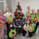 Siedem dziewczynek pozuje przy choince ozdobionej światełkami i ozdobami. Każde dziecko trzyma własnoręcznie wykonany z papieru wieniec z zielonymi liśćmi, czerwoną kokardą i świecą. W tle białe ściany, okno z cienkimi, białymi firankami i biały stół z komputerem.